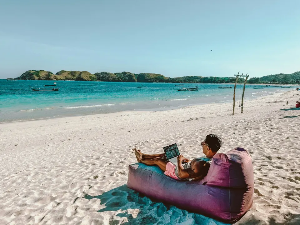 Pessoa deitada numa almofada na areia, com o notebook, numa praia de águas turquesa
